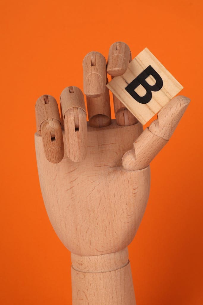 A realistic wooden mannequin hand holding a letter B block against an orange background.