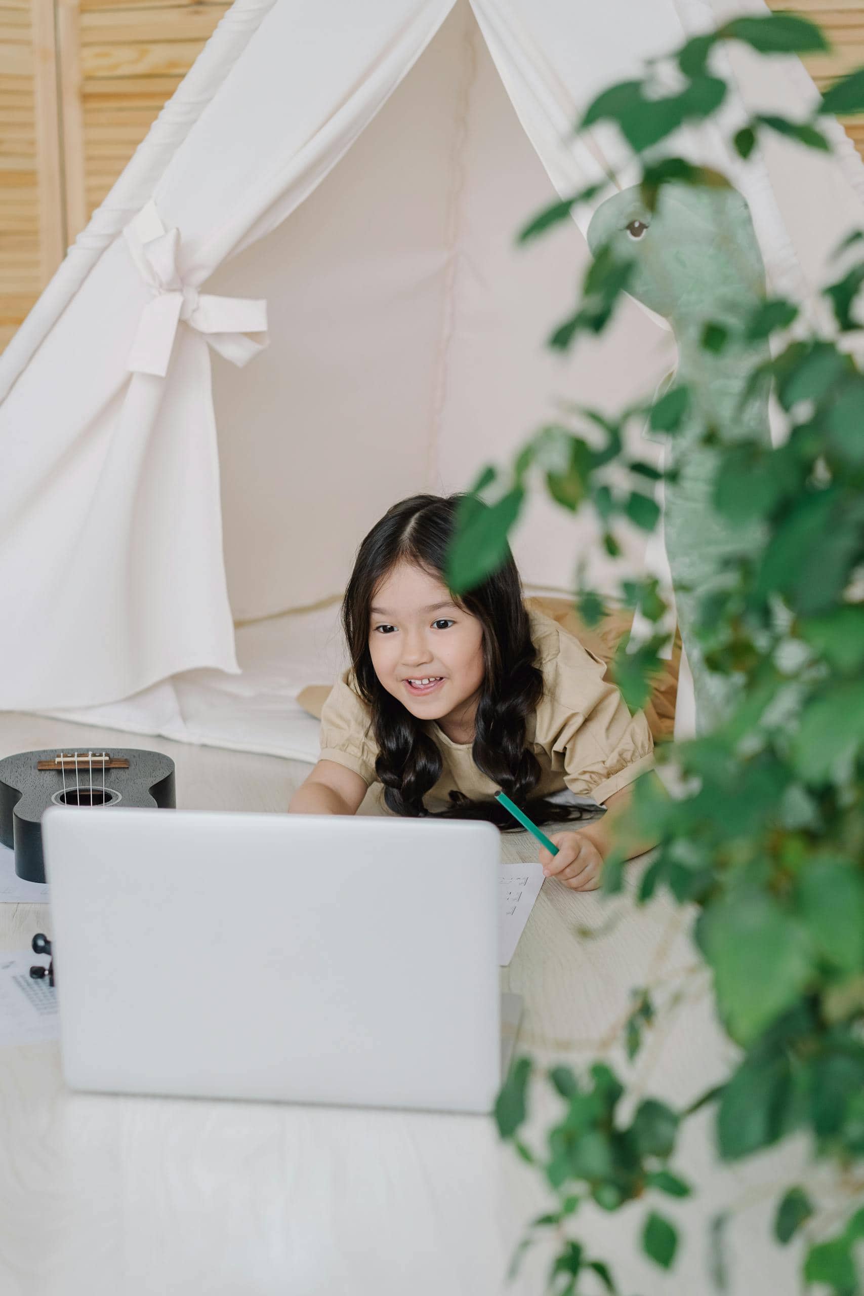 A young girl smiles while interacting with a laptop inside a cozy indoor tent, surrounded by a relaxing home setting.